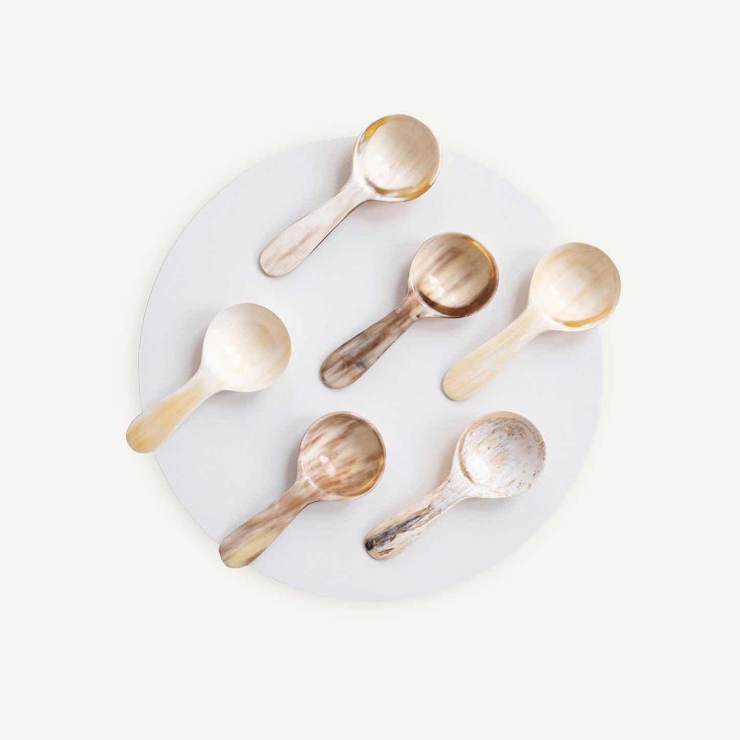 Six hand-carved horn coffee scoops with varied natural coloring and grain, arranged on a round plate, viewed from above